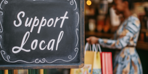 Photo of a sign that reads Support Local on the front of a small business. A woman is shopping in the background.