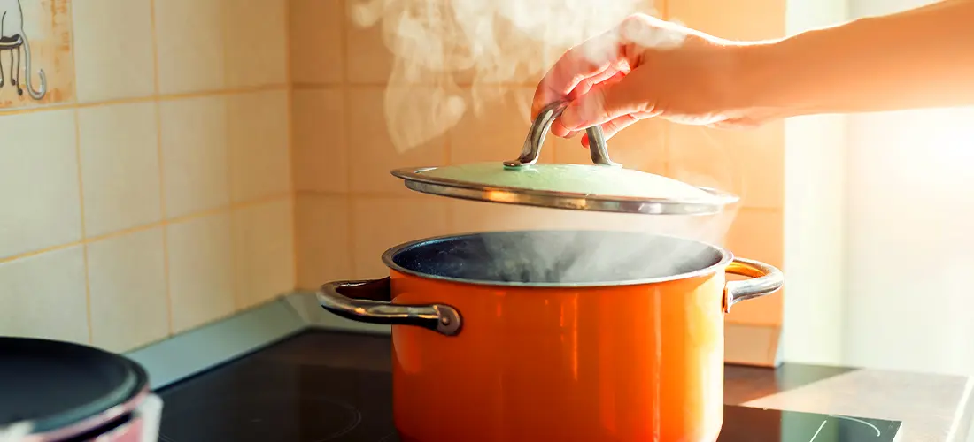 An orange pot of boiling water on the stove, the top being lifted off the pot to show steam rising from the pot of boiling water.